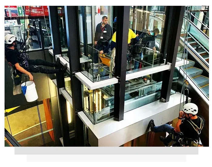 Elevator maintenance: workers rappelling inside glass shaft, one holding a bucket, another on a platform, escalators in the background.