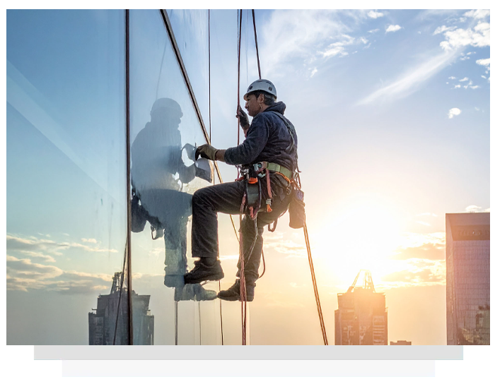 Window cleaner suspended from ropes, cleaning a tall skyscraper with the setting sun in the background.