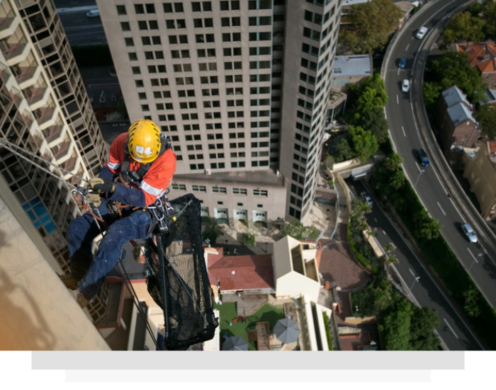 Worker in yellow helmet rappelling beside a tall building, with city streets and apartments below.