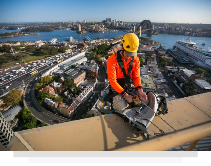 Worker in orange safety gear on a rooftop, using a tool with a city and harbor skyline behind him