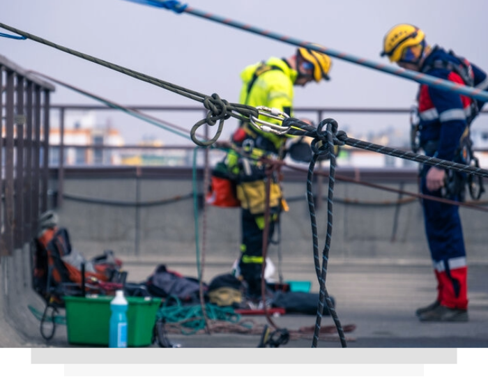 Rope technician in orange helmet and harness rappelling beside a sloped rooftop.