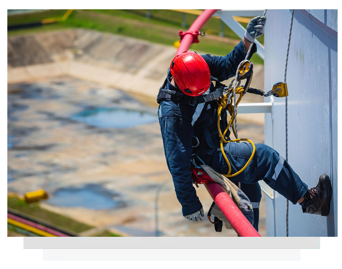 Worker in safety gear on a tall structure, securing a red hose, with industrial setting in background.