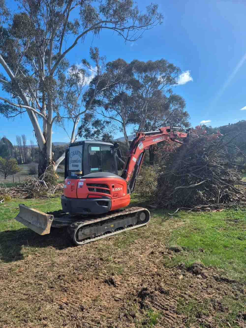 A Small Excavator Is Sitting In A Field Next To A Pile Of Branches — Grants Excavations In Osbornes Flat, VIC