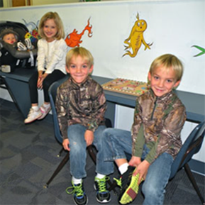 Three children are sitting in chairs in front of a wall with drawings on it