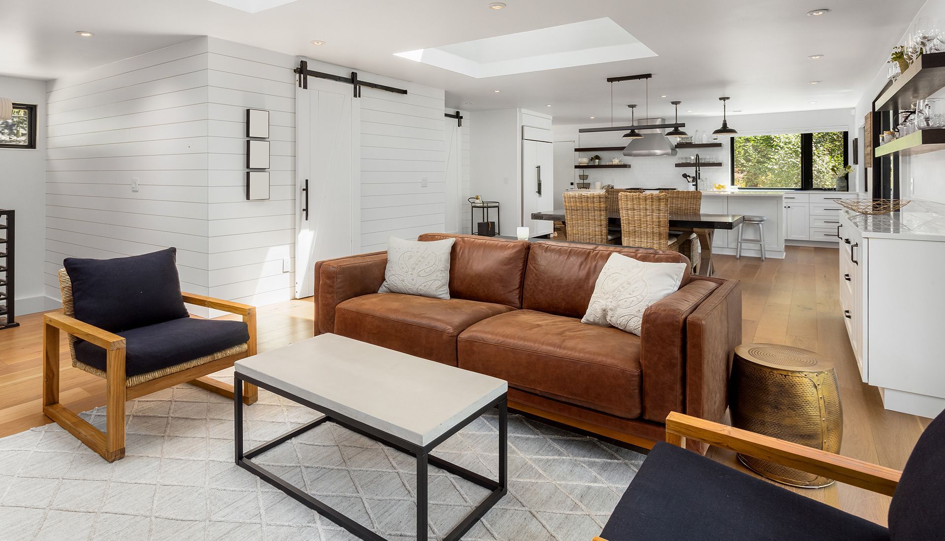 A bright, modern living room features a tan leather sofa, two navy accent chairs, a concrete coffee table, and a kitchen.