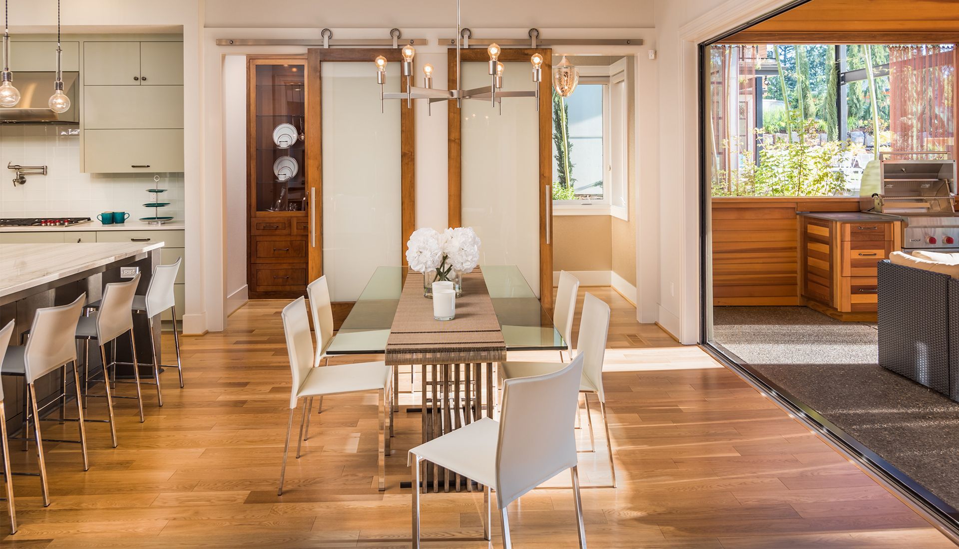 A bright open-concept dining room with a glass table, modern white chairs, and sliding doors opening to a patio.