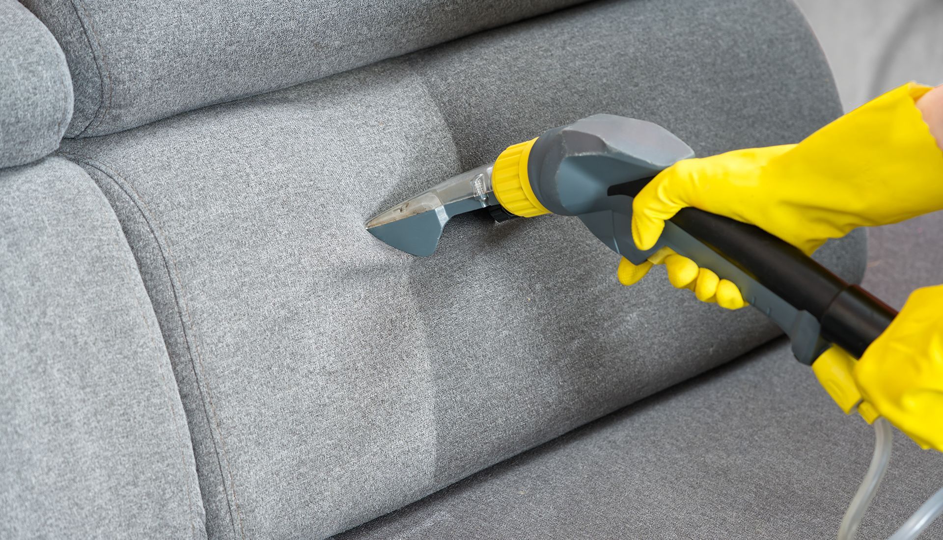 A person wearing yellow rubber gloves uses a steam cleaner tool to deep clean a grey fabric sofa.