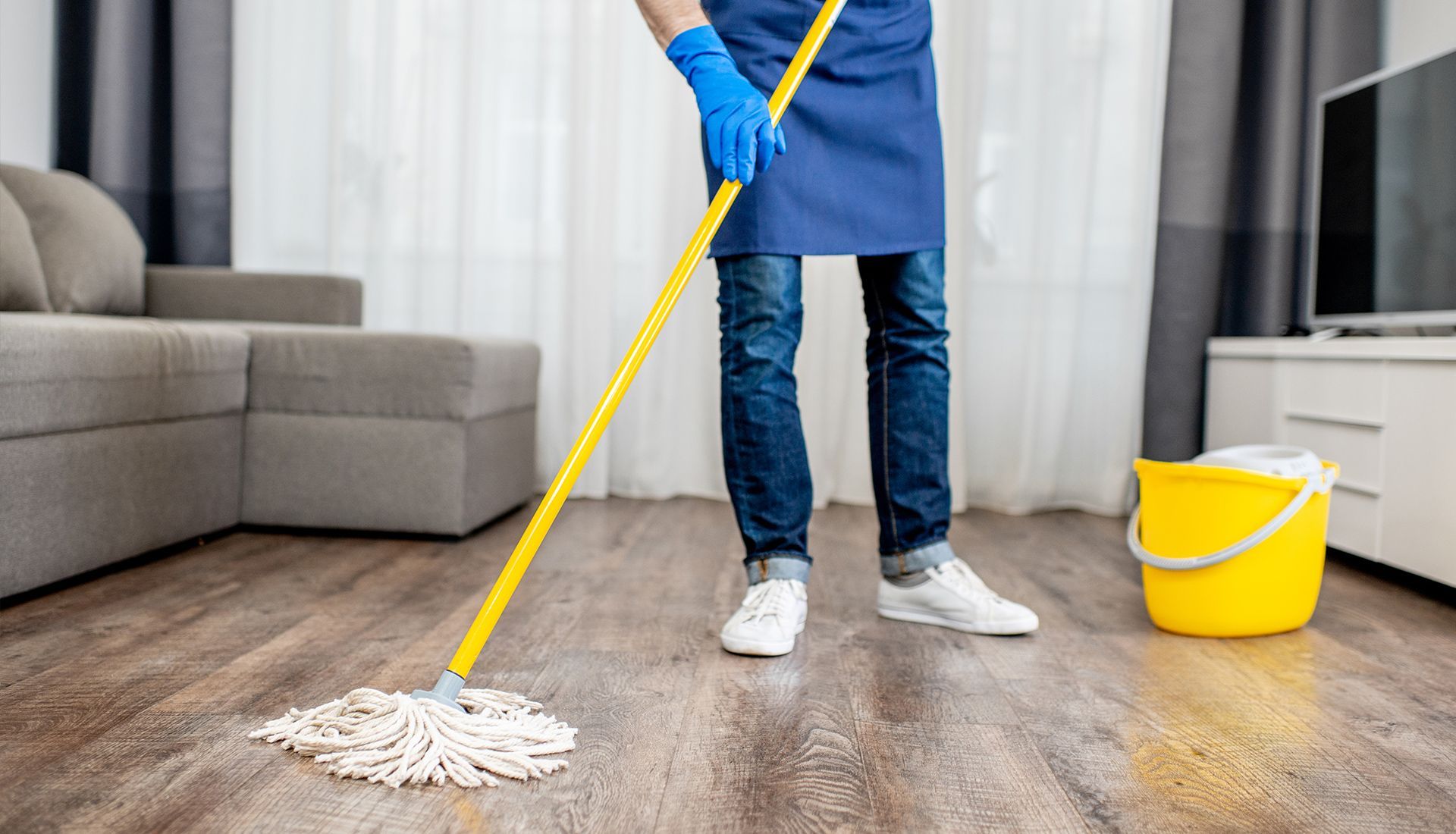 A person wearing a blue apron and gloves mops a wooden floor in a living room near a yellow bucket.