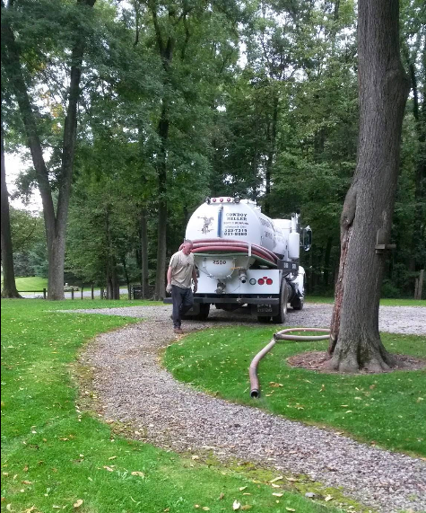 Worker cleaning a white truck on a winding gravel path in a grassy park with trees