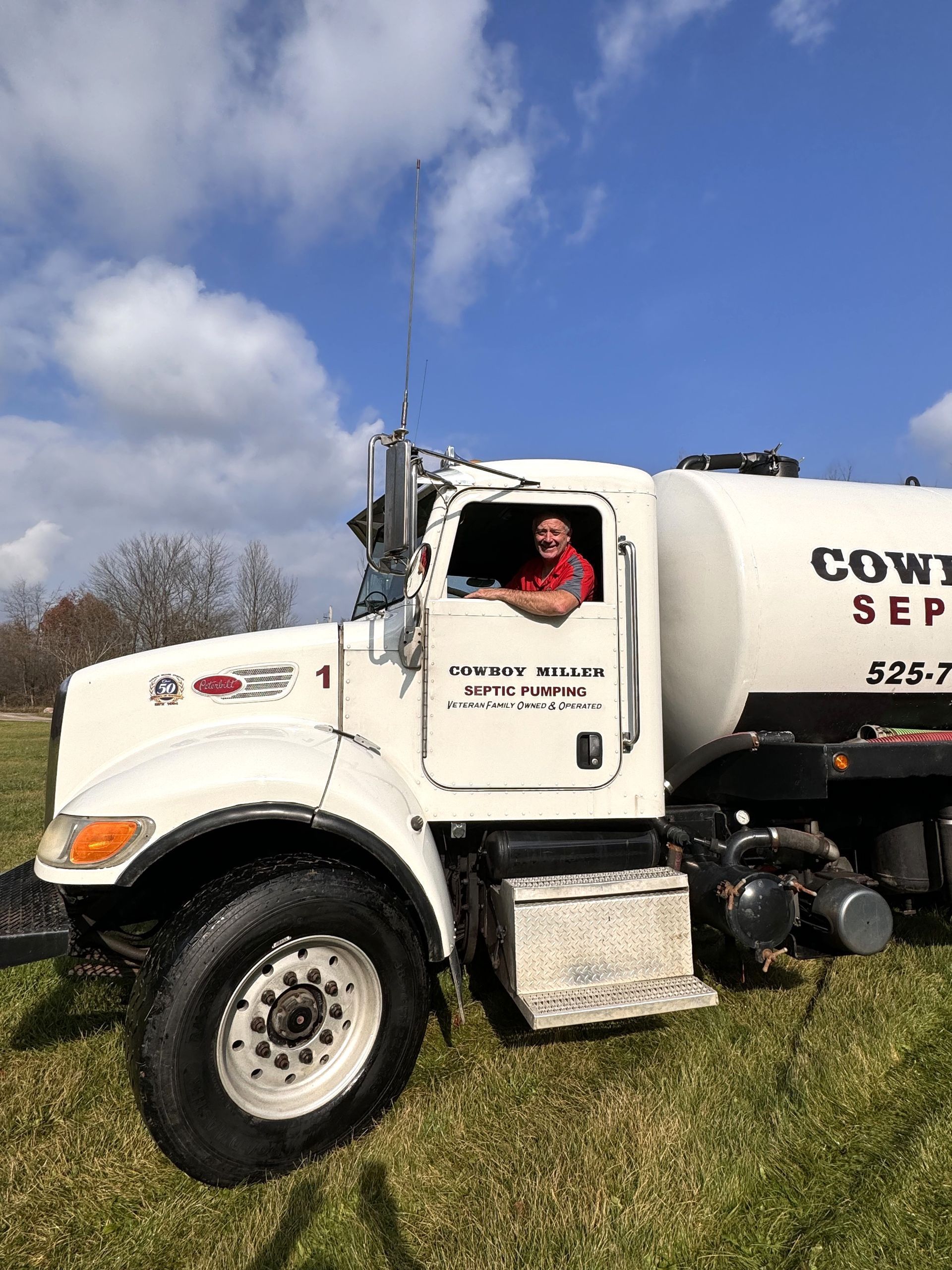 White tanker truck parked on grass under a blue sky, with a driver visible in the cab.