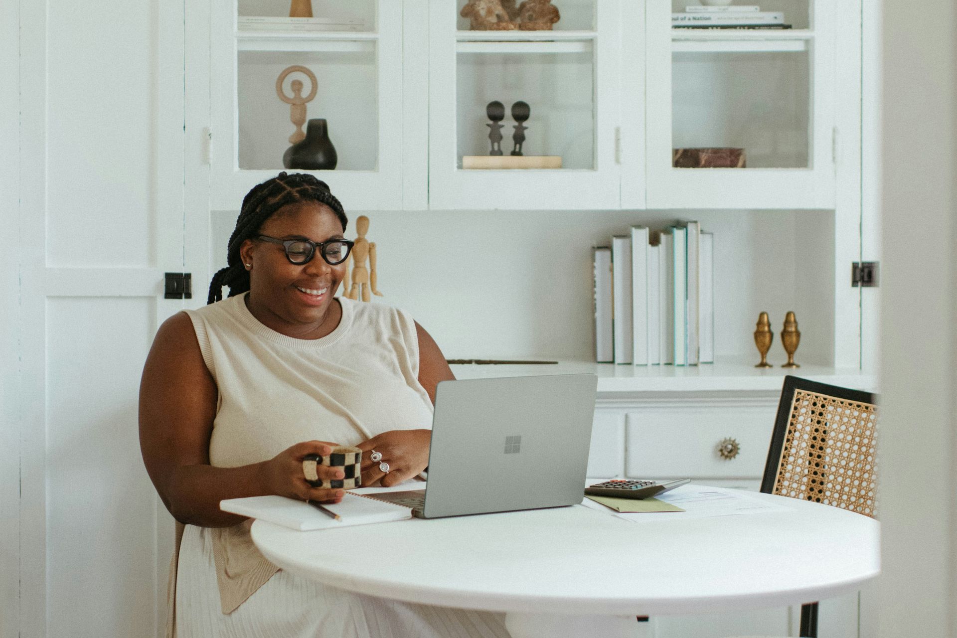Woman at a white table, smiling at a laptop. White built-in shelves behind her.