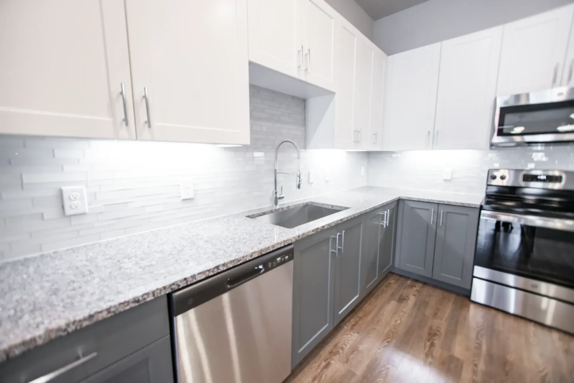 Modern apartment kitchen with white upper cabinets, gray lower cabinets, granite counters, and stainless appliances.