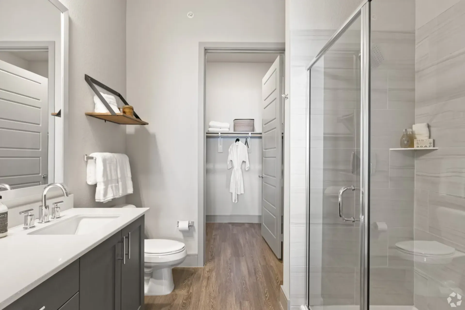 Modern bathroom with a double-sink vanity, toilet, glass-enclosed shower, and a walk-in closet visible through an open doorway.