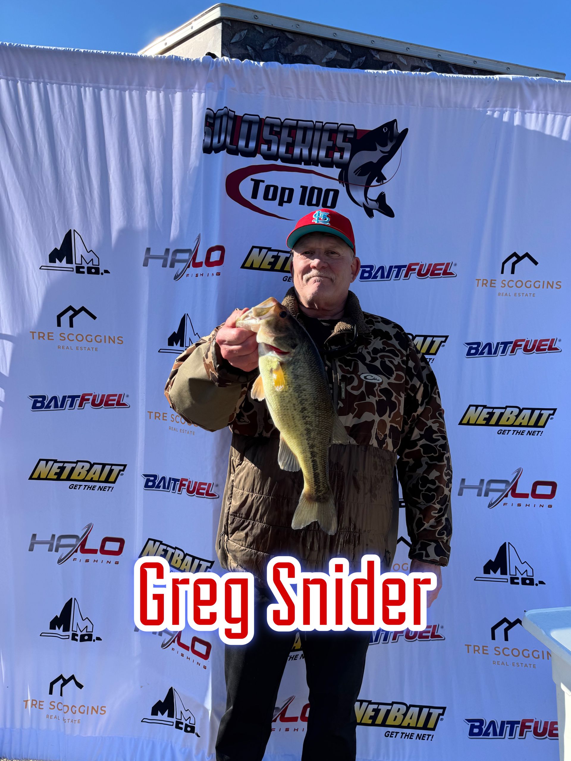 Man holding a large fish at an outdoor event backdrop with sponsor logos and the name “Greg Snider”