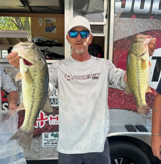 Angler holding a trophy and check at a fishing tournament podium, wearing a sponsor jersey and cap.