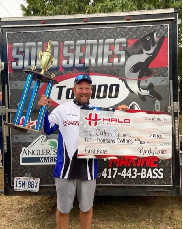 Man standing on a sponsor backdrop, wearing a white fishing jersey and cap, holding a trophy plaque.