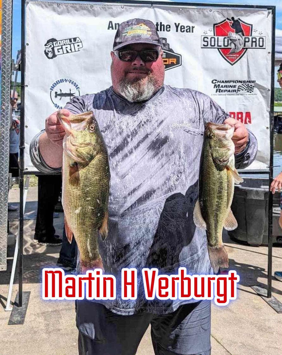 Man holding two large bass fish in front of a sponsor banner, with “Martin H Verburg” text.