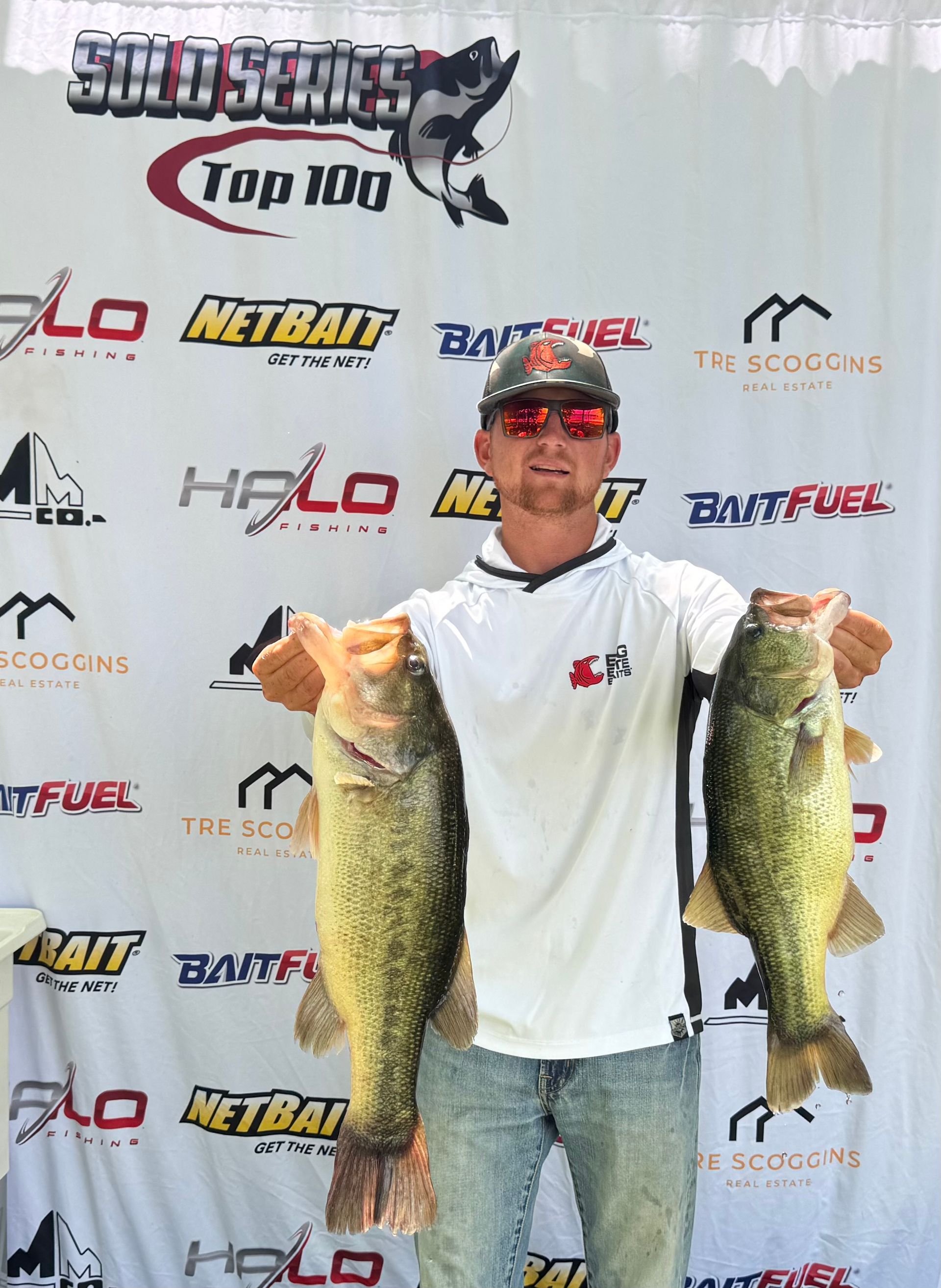 Angler holding two large fish in front of a sponsor backdrop at a fishing event