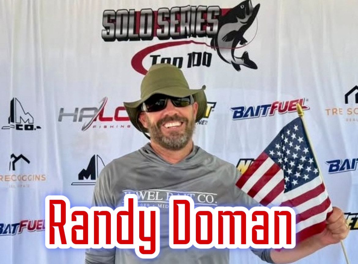 Randy Doman smiling in sunglasses and cap, holding a small American flag at a racing event backdrop
