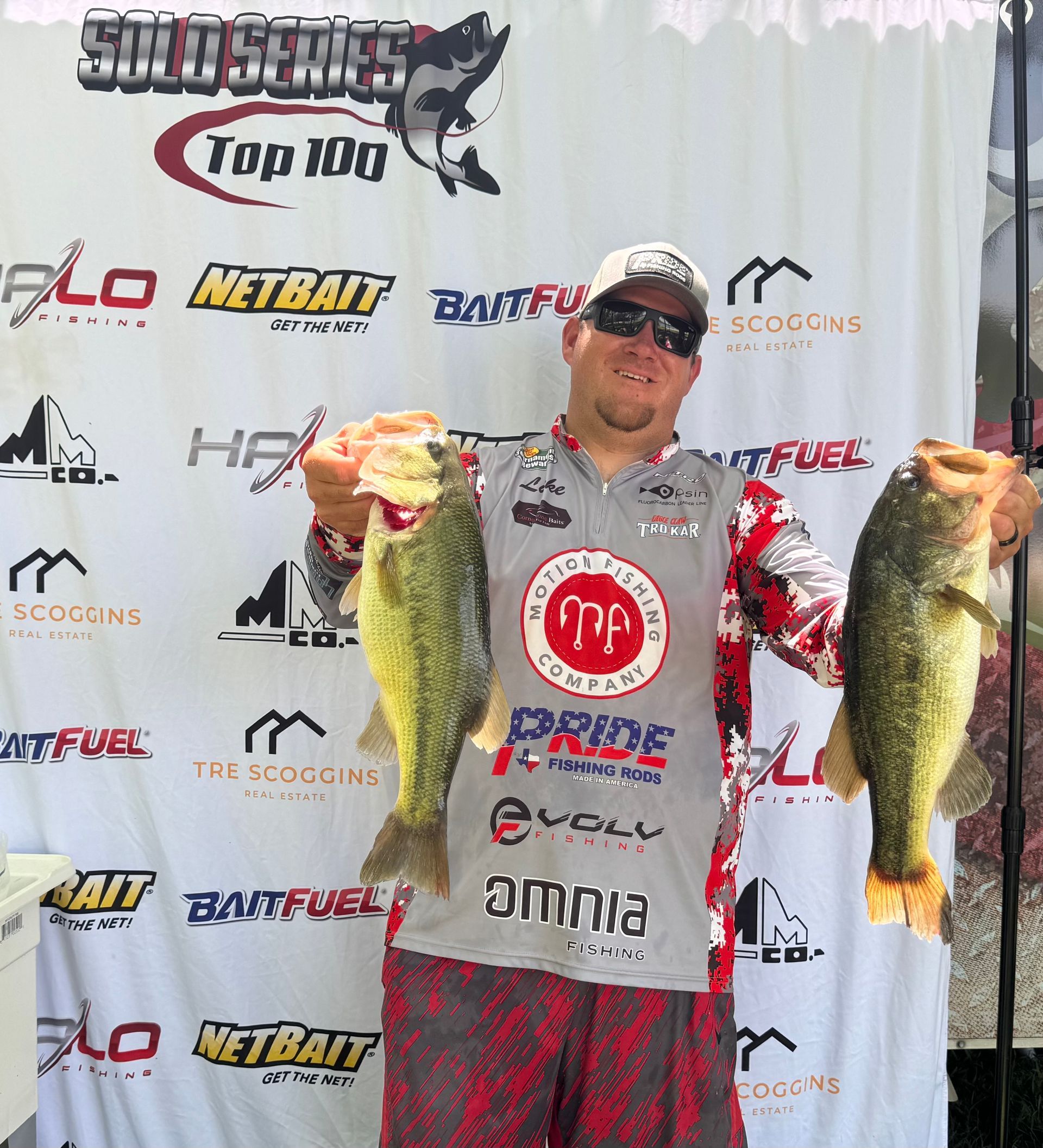 Angler holding two large bass at a fishing tournament backdrop, wearing a cap and sponsor jersey.