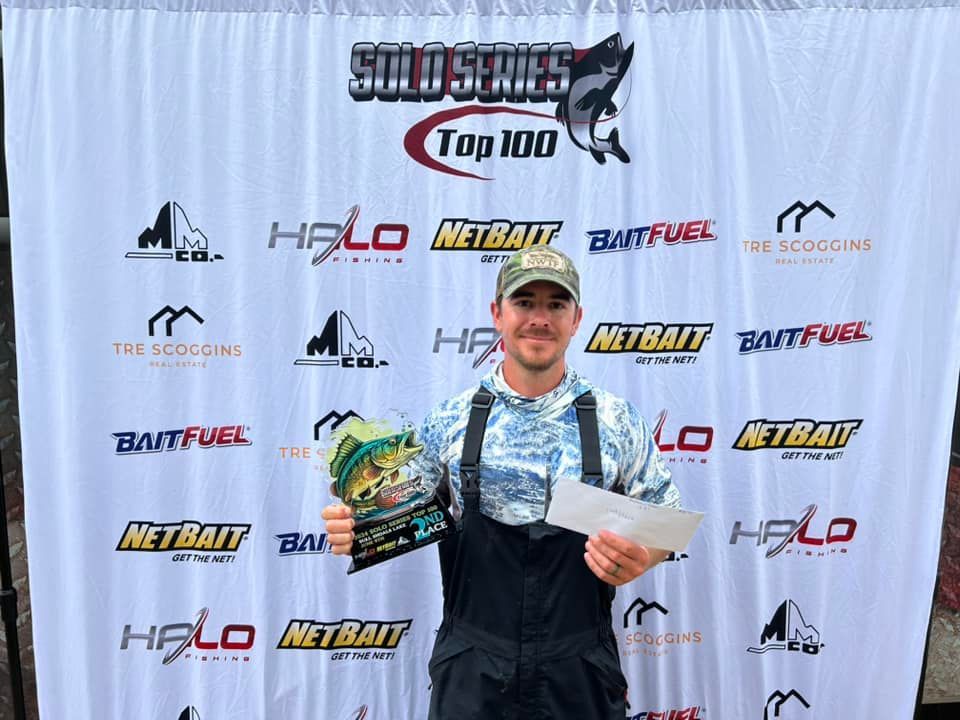 Man holding two trophies and a prize bag at a fishing tournament backdrop, smiling indoors