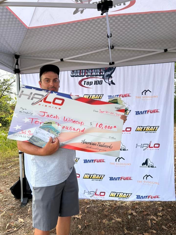 Man holding a large fish trophy under a canopy at a fishing event, with sponsor banners behind him.