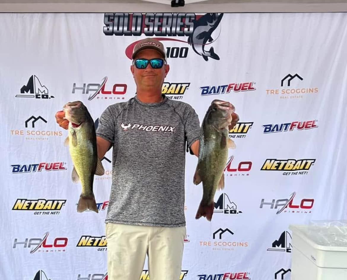Man holding two large fish in front of a fishing tournament backdrop