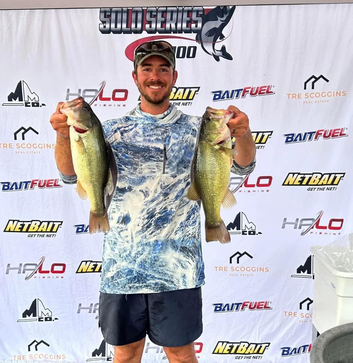 Angler smiling and holding two largemouth bass at a tournament backdrop