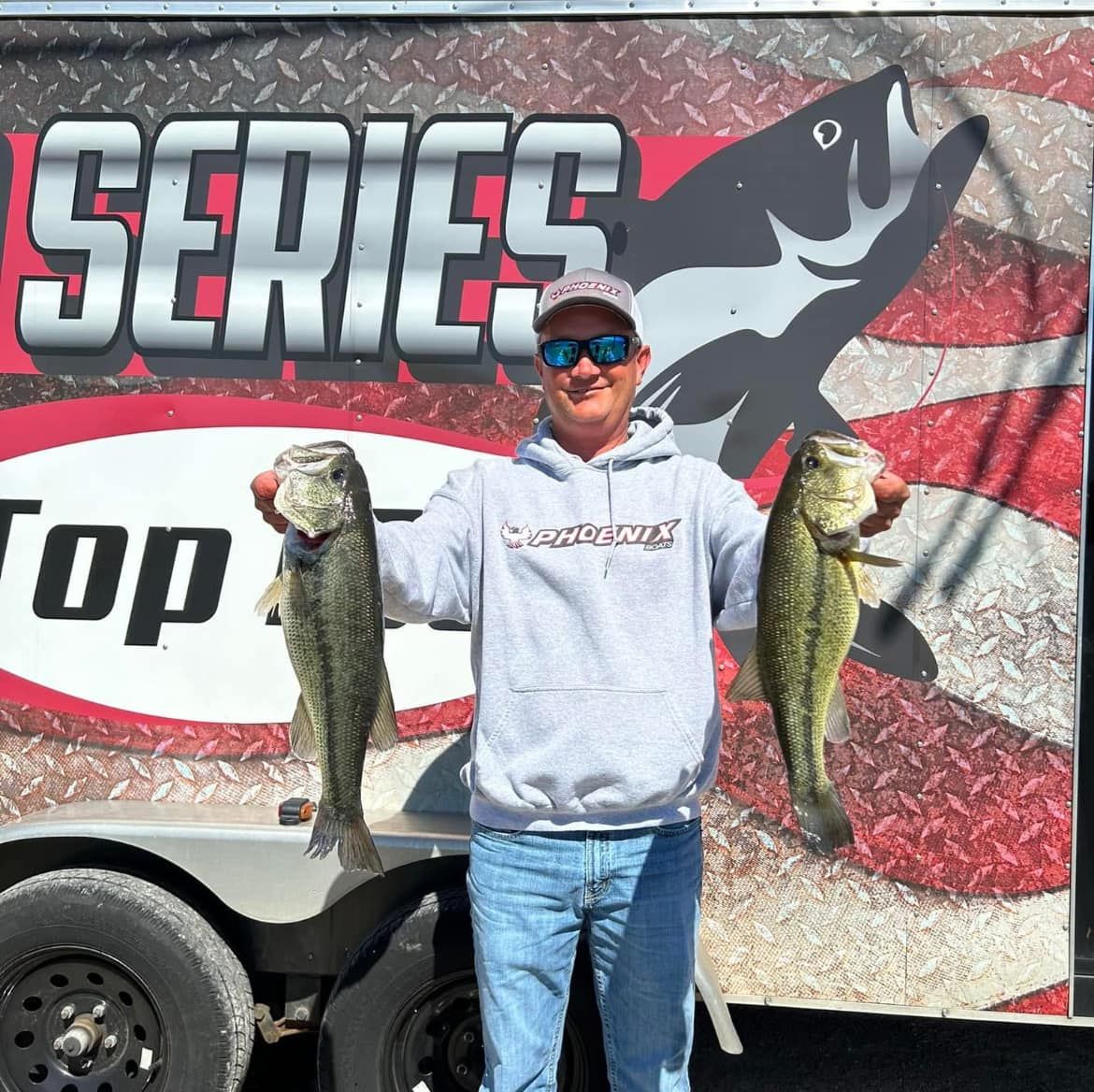 Man holding two largemouth bass in front of a fishing trailer with a “SERIES” banner.