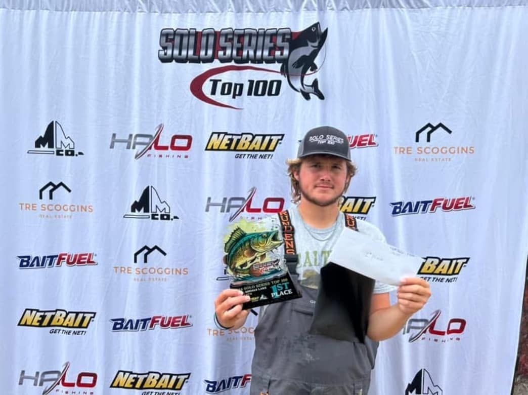 Person holding a trophy and certificate at a sports awards backdrop, smiling in a cap and gray shirt