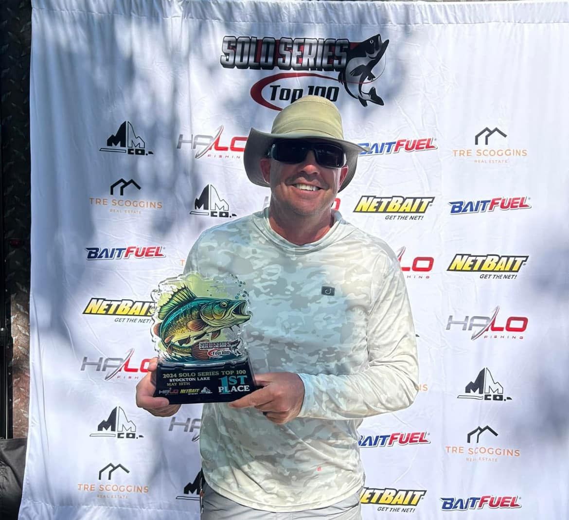 Smiling person in white shirt and cap holds a trophy in front of a sponsor backdrop.
