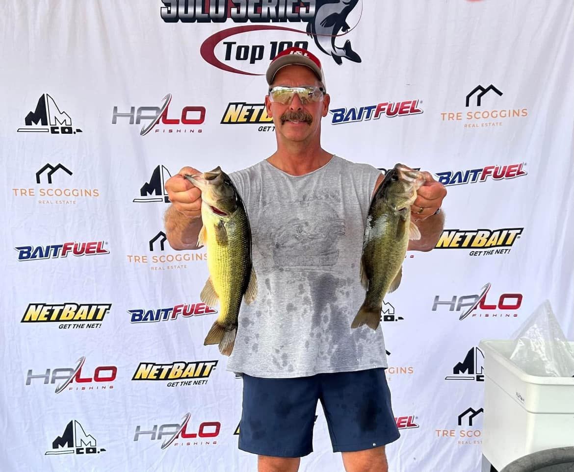 Man holding two large fish in front of a fishing tournament sponsor backdrop