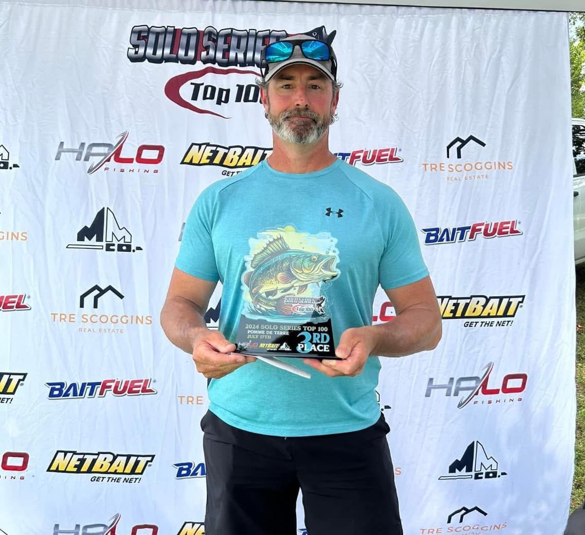 Man holding a trophy in front of a sponsor backdrop, wearing a turquoise shirt and cap.