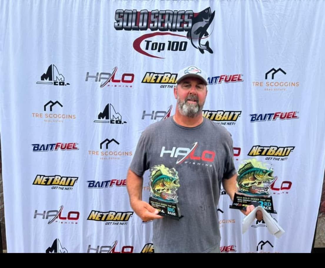 Man in gray shirt and cap holding two trophies at a fishing tournament awards backdrop