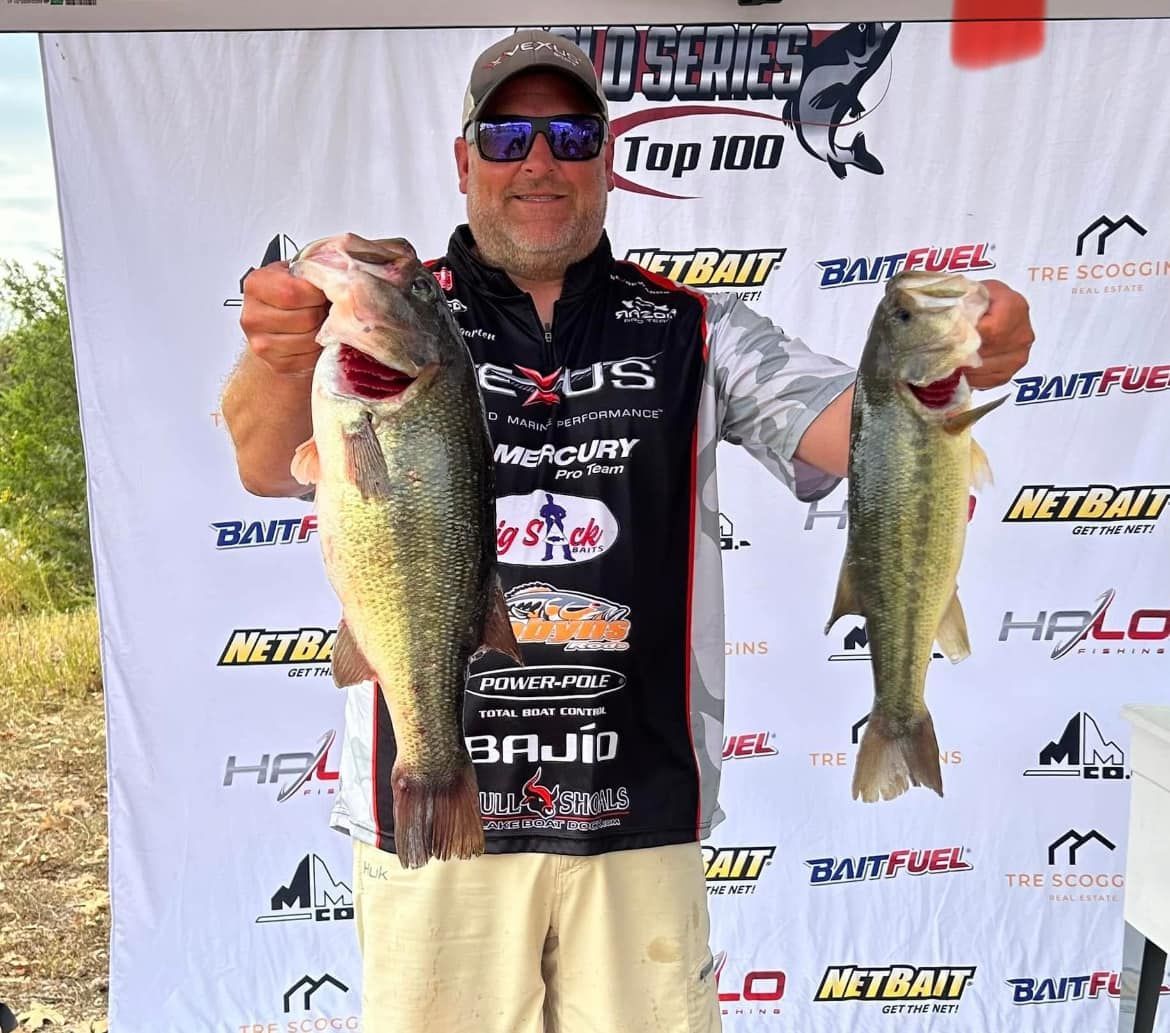 Man holding two largemouth bass in front of a fishing tournament backdrop, wearing sunglasses and a sponsor jersey