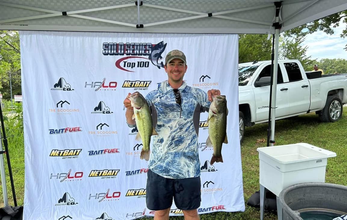 Angler under a canopy holding two largemouth bass at an outdoor fishing tournament