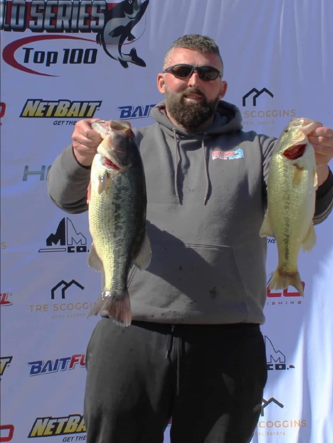 Man holding two large fish in front of a fishing tournament backdrop.