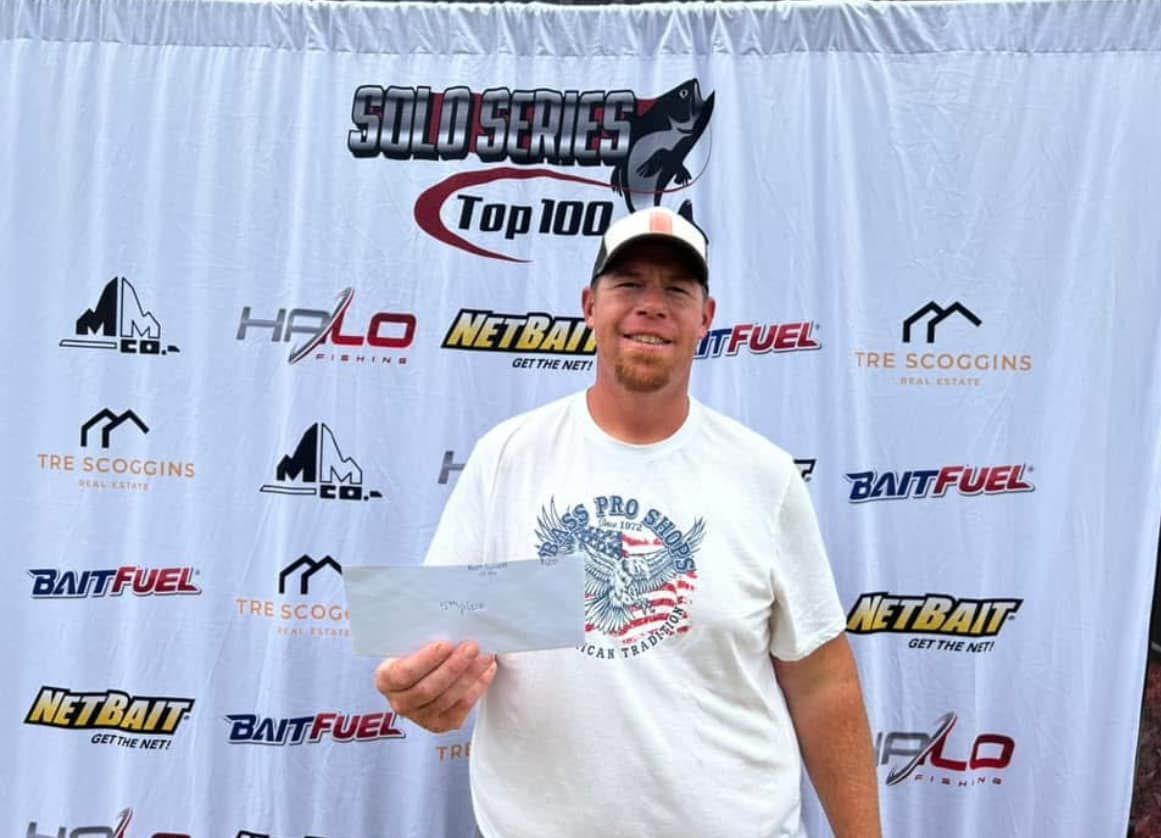 Man holding a fish in front of a branded fishing tournament backdrop, wearing a white T-shirt and cap.