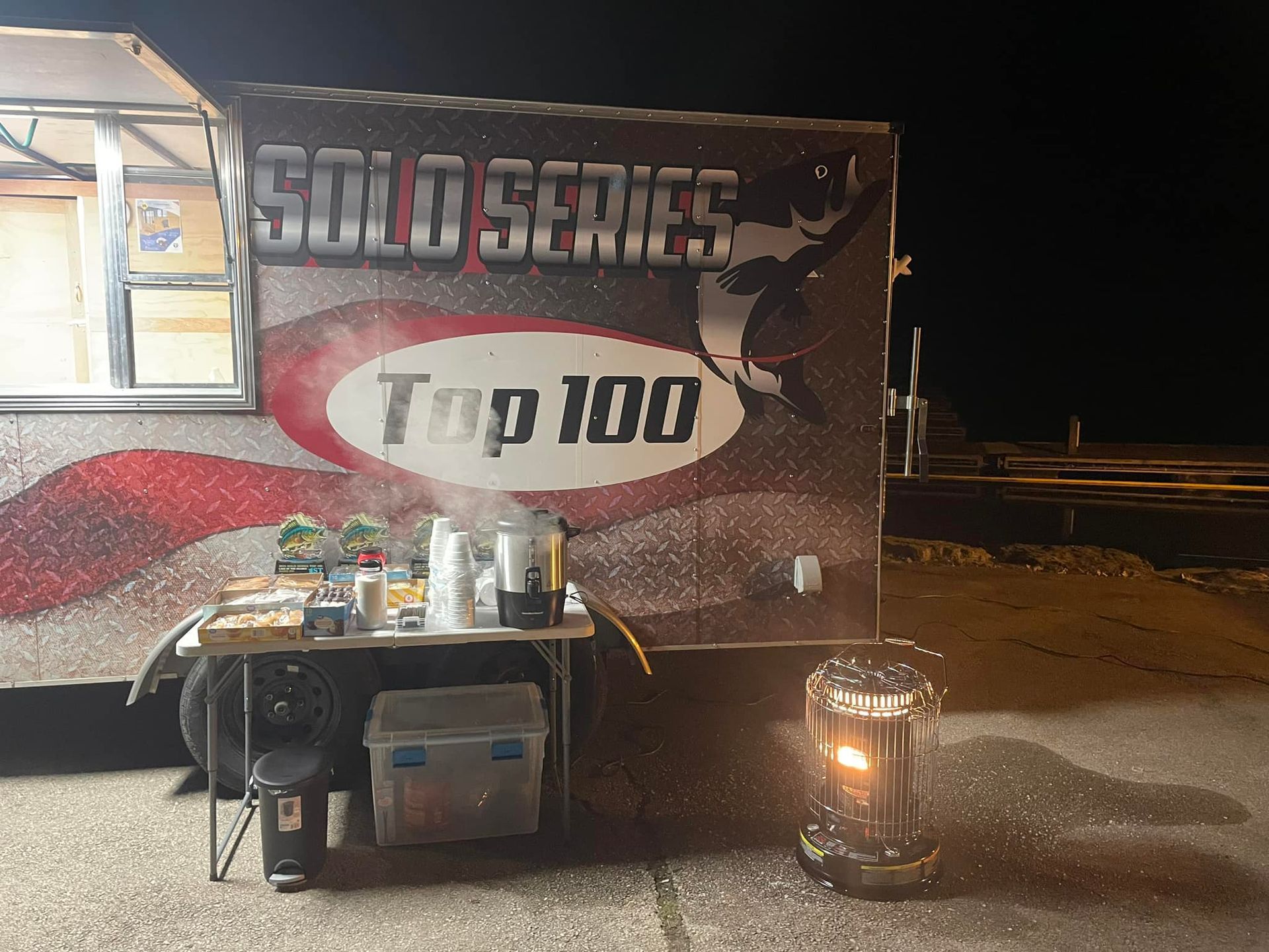 Nighttime race trailer with “SULI SERIES Top 100” banner, table of drinks, and a propane heater nearby