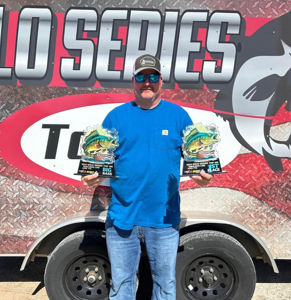 Man in blue shirt holding two trophies in front of a trailer with bold racing-style graphics.