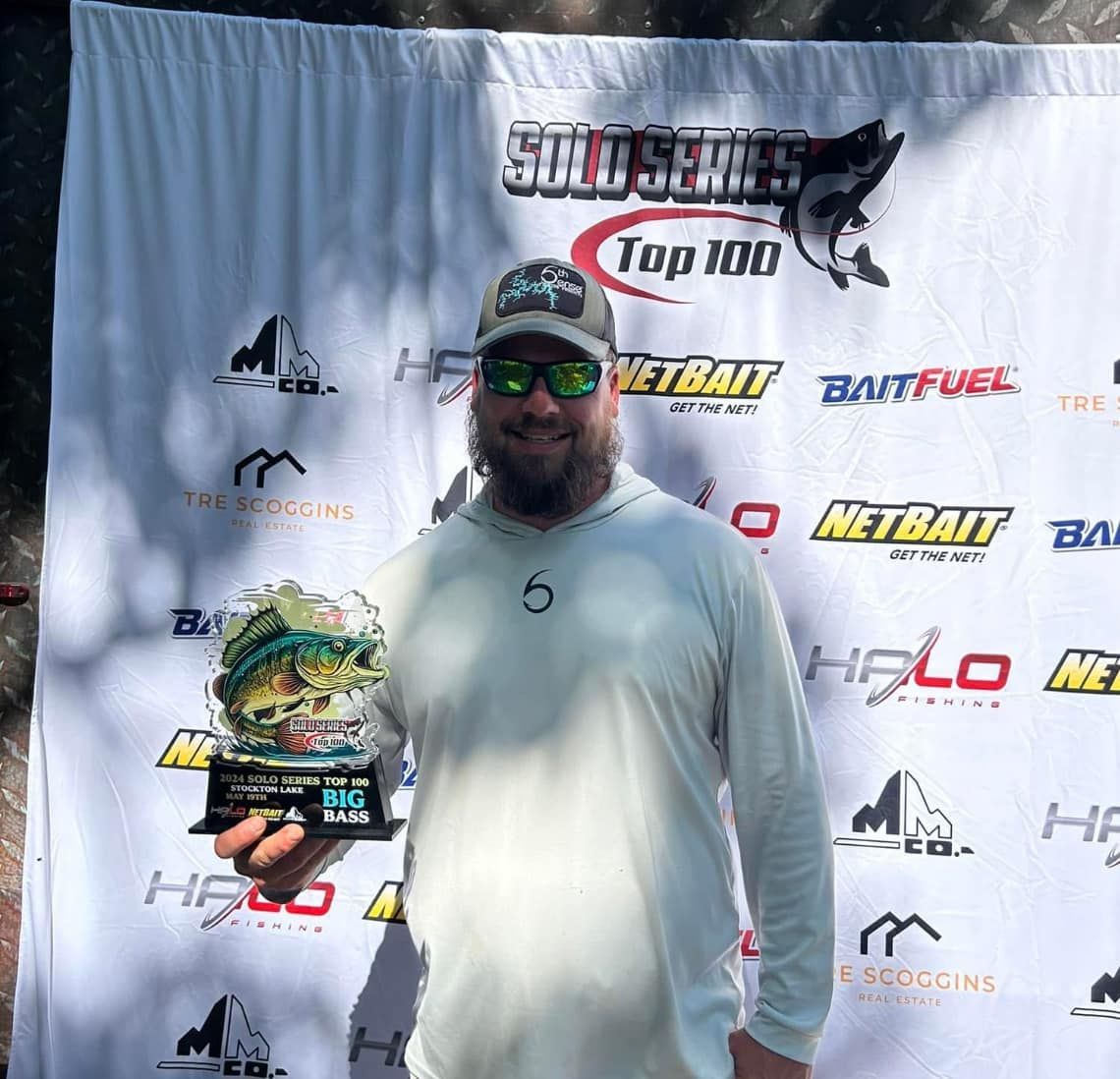 Man holding a trophy in front of a sponsor banner at an outdoor awards event