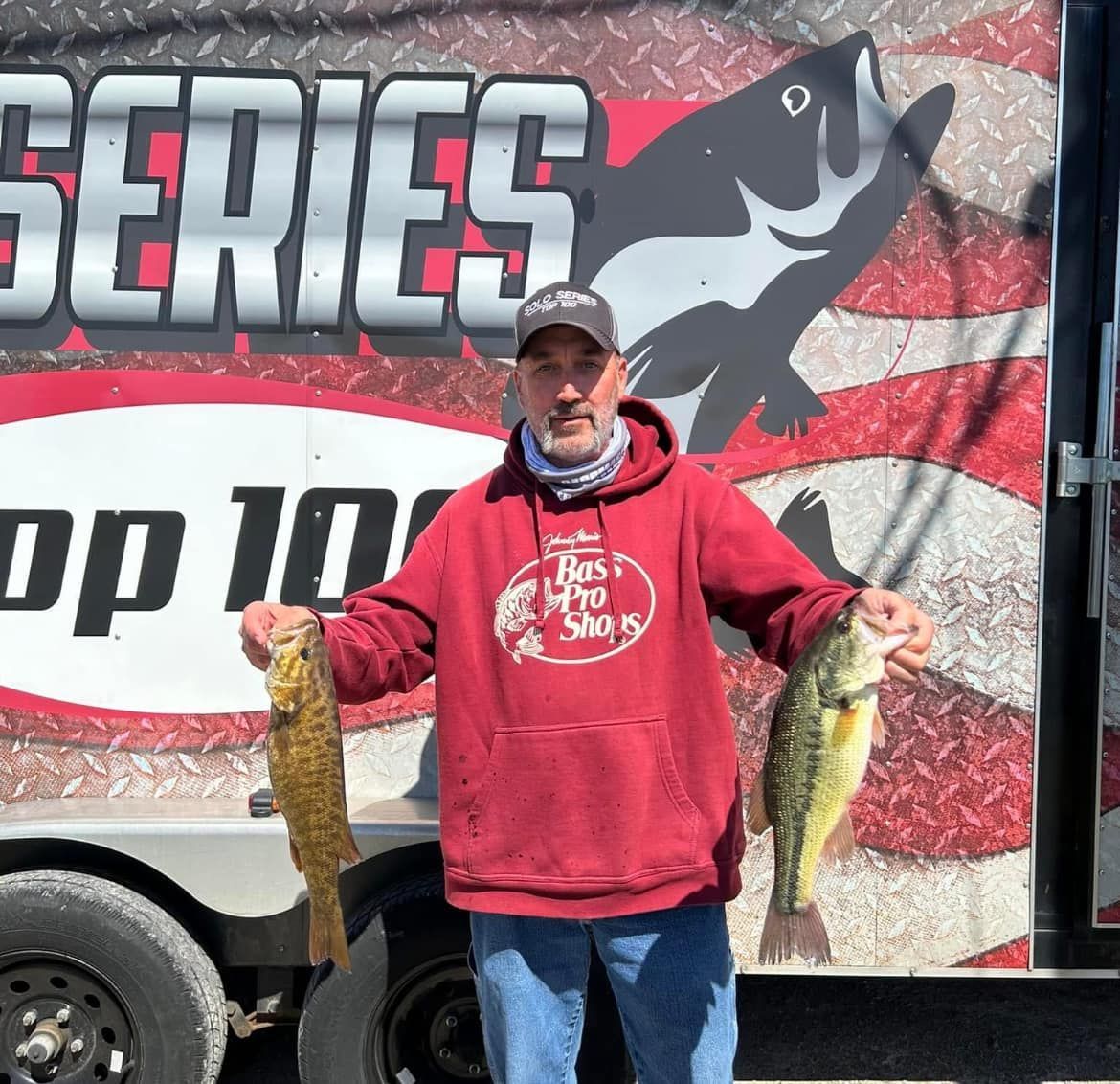 Man in a red hoodie holding two large fish in front of a fishing tournament banner.