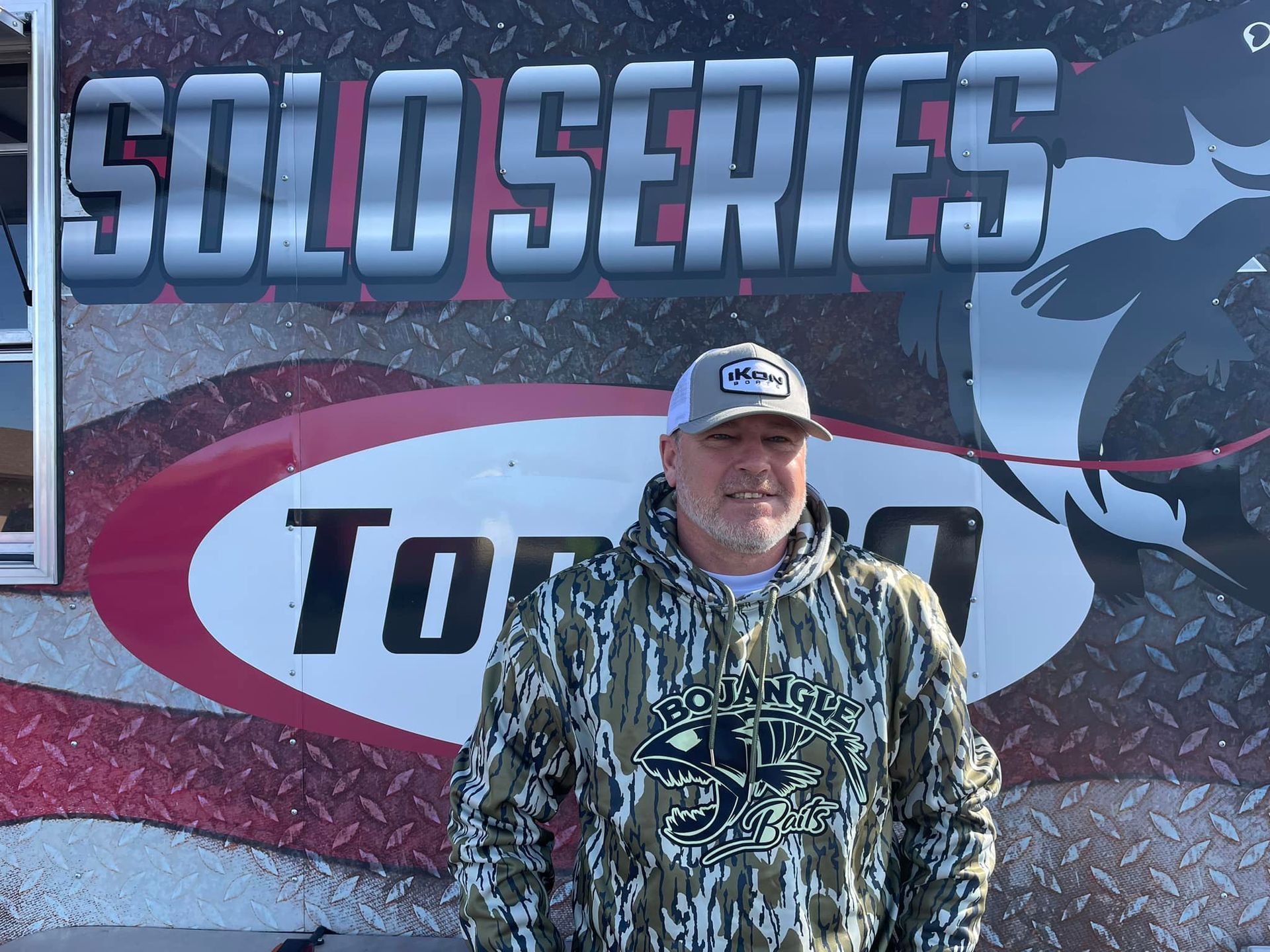 Man in camouflage hoodie standing before a Solo Series race banner and truck logo