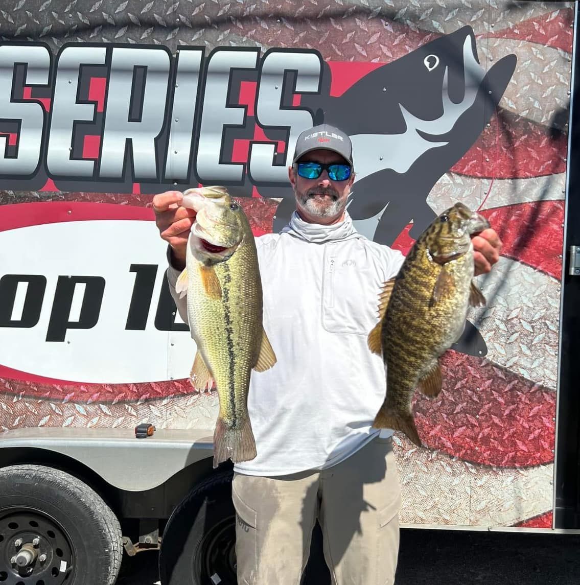 Man holding two large fish in front of a fishing tournament banner and trailer.
