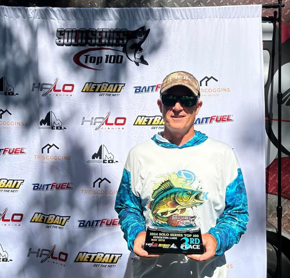 Person holding a trophy in front of a sponsor banner at an outdoor fishing event.