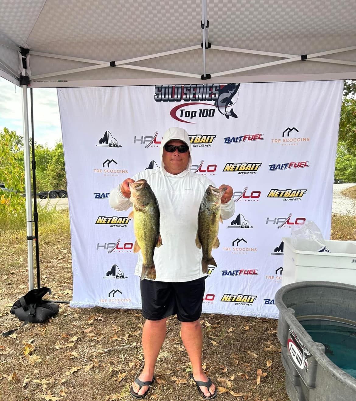 Angler holding two large fish in front of a fishing tournament backdrop under a canopy.