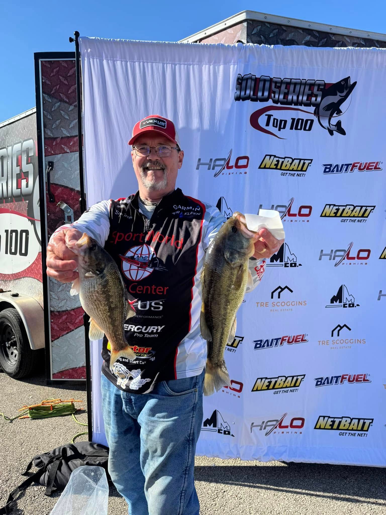 Angler holding two large fish in front of a sponsor backdrop at an outdoor fishing event