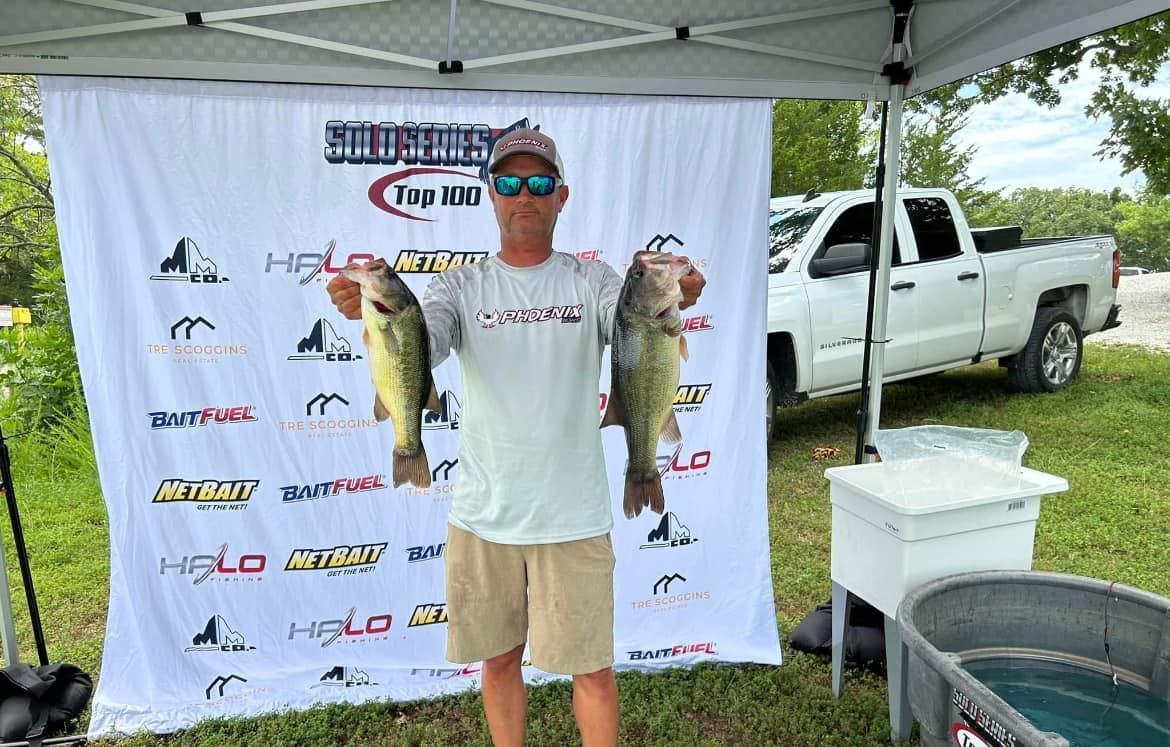 Man holding a large fish under a canopy beside a pickup truck and event banner