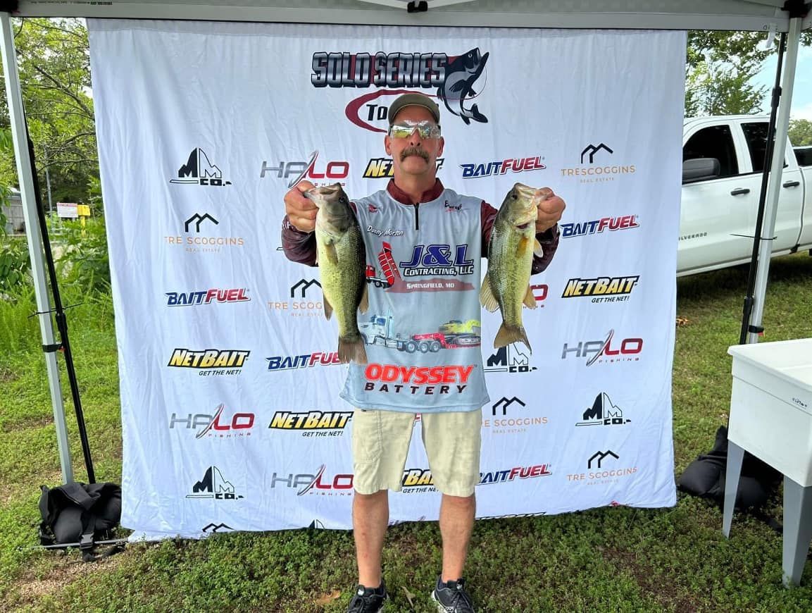 Angler holding two bass in front of a sponsor banner at an outdoor fishing event