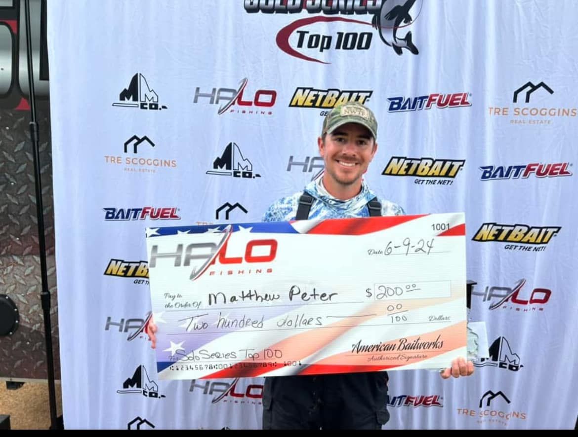 Man holding a giant check at a race event podium with sponsor banners behind him